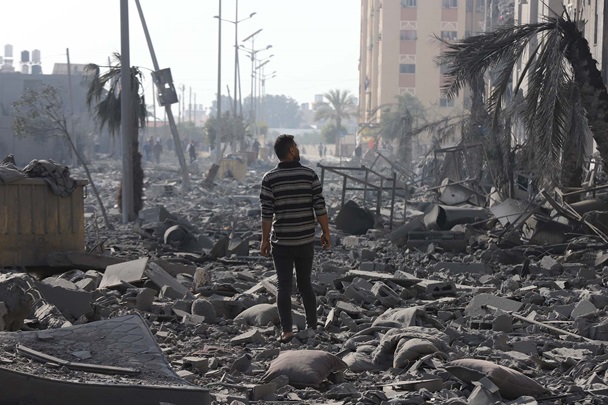 En mann sitter med hodet i hendene foran en utbombet blokk med leiligheter.