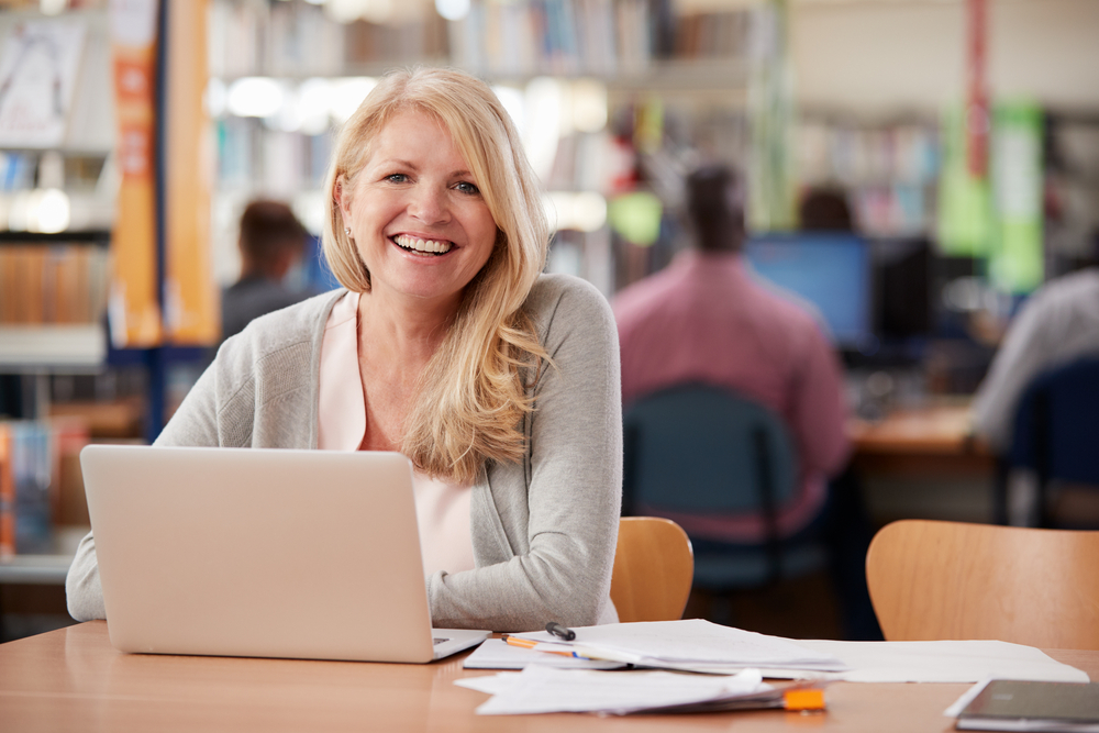 Kvinne smiler mot kamera med en laptop foran seg. I bakgrunnen ser man flere studenter som sitter rundt maskiner. Bilde.