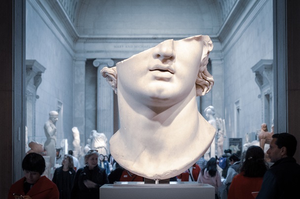 Gresk skulptur i forgrunnen som viser deler av hode i bakgrunnen en gruppe mennesker som er på guided tur i et museum