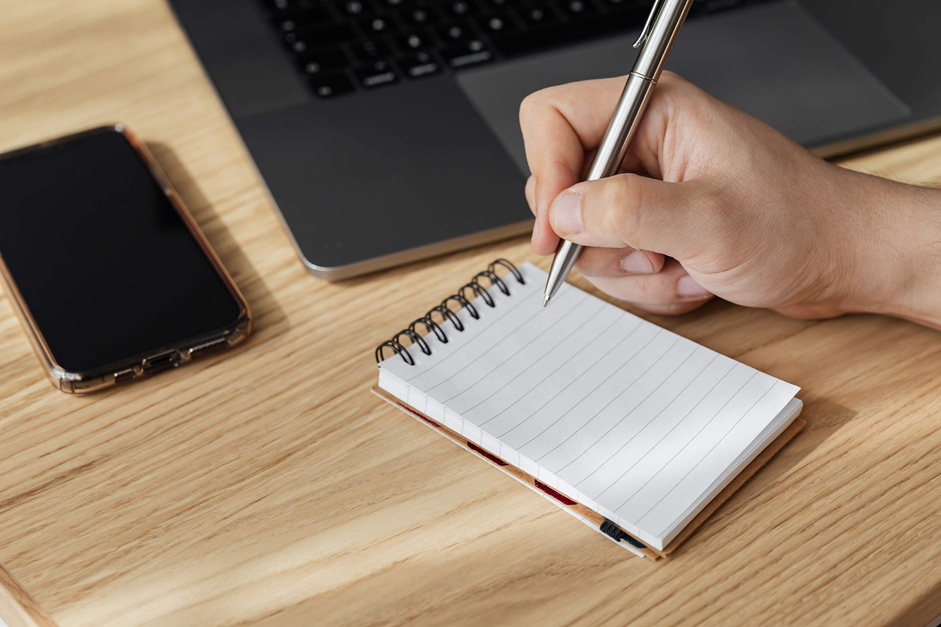 En hånd skriver på en notatblokk foran en laptop og en mobil