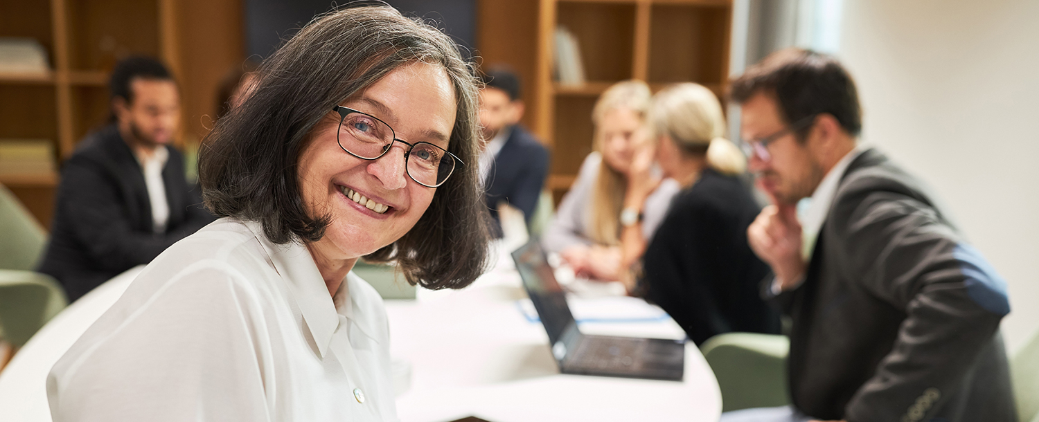 En eldre kvinne smiler til kamera ved et møtebord fullt av kolleger som jobber