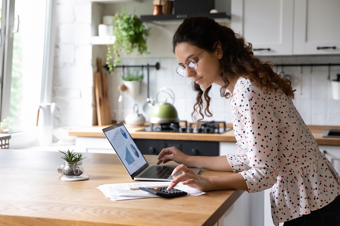 Kvinne i profil som står ved et kjøkkenbord med en PC og kalkulator foran seg