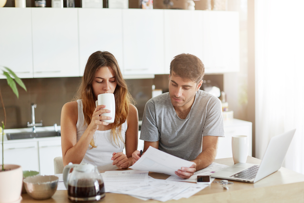 Kvinne og mann ser på papirer i et kjøkken, kvinnen drikker kaffe.