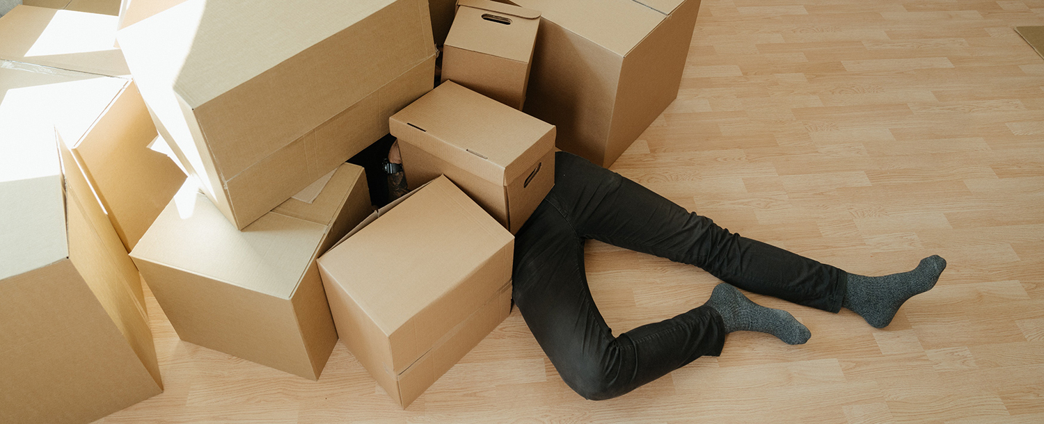 En mann ligger på gulvet og bena stikker ut fra en haug med pappesker. 