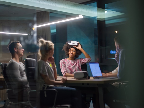 Fire personer sitter i et møterom og den ene møtedeltakeren holder et VR-sett mot håret. Det er også et VR-sett og pc på bordet.  
