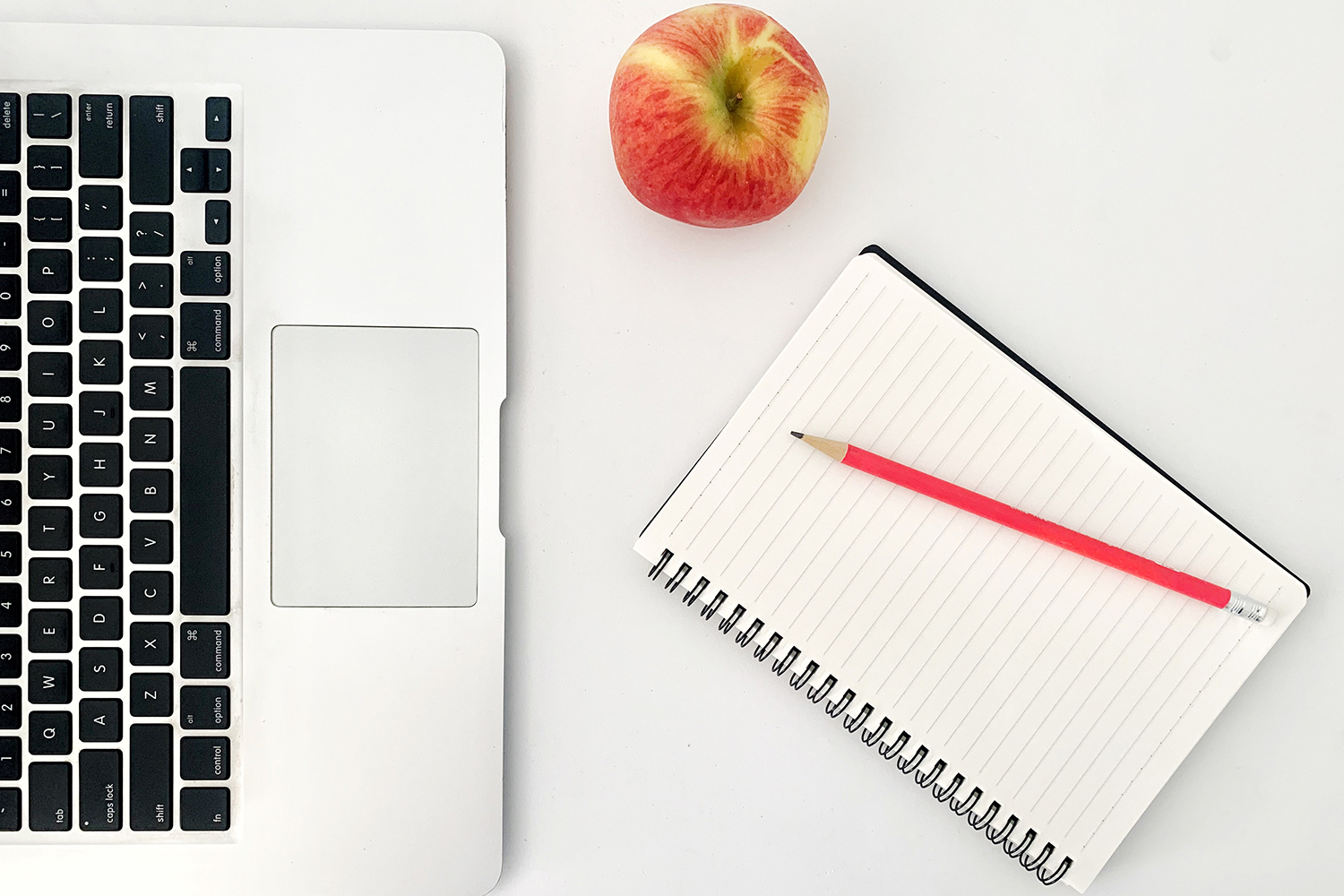 Et skrivebord med en mac, et eple, en skrivebok og en rød penn. 
