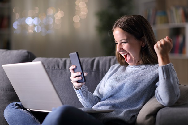 En kvinne sitter i sofaen og jubler mens hun ser på mobilen. På fanget har hun en pc