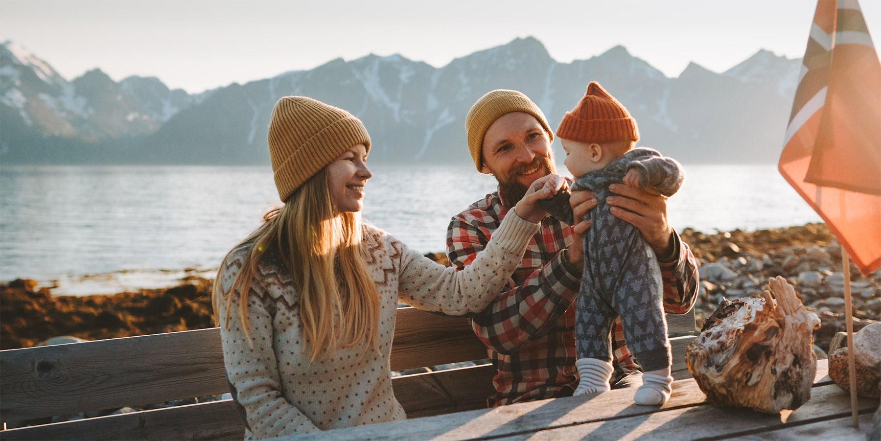 Mor, far og et lite barn sitter ved et trebord langs vannkanten i et idyllisk norsk fjordlandskap